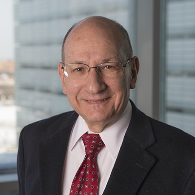 Dr. Coleman R. Seskind smiles in a suit and red tie against a blurred skyscraper background