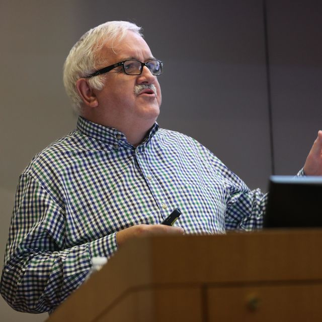 Professor Sir Stephen O&#039;Rahilly stands behind a wooden podium talking while presenting to an audience out of camera view. His left hand is outstretched and his right hand is holding a remote at chest level.